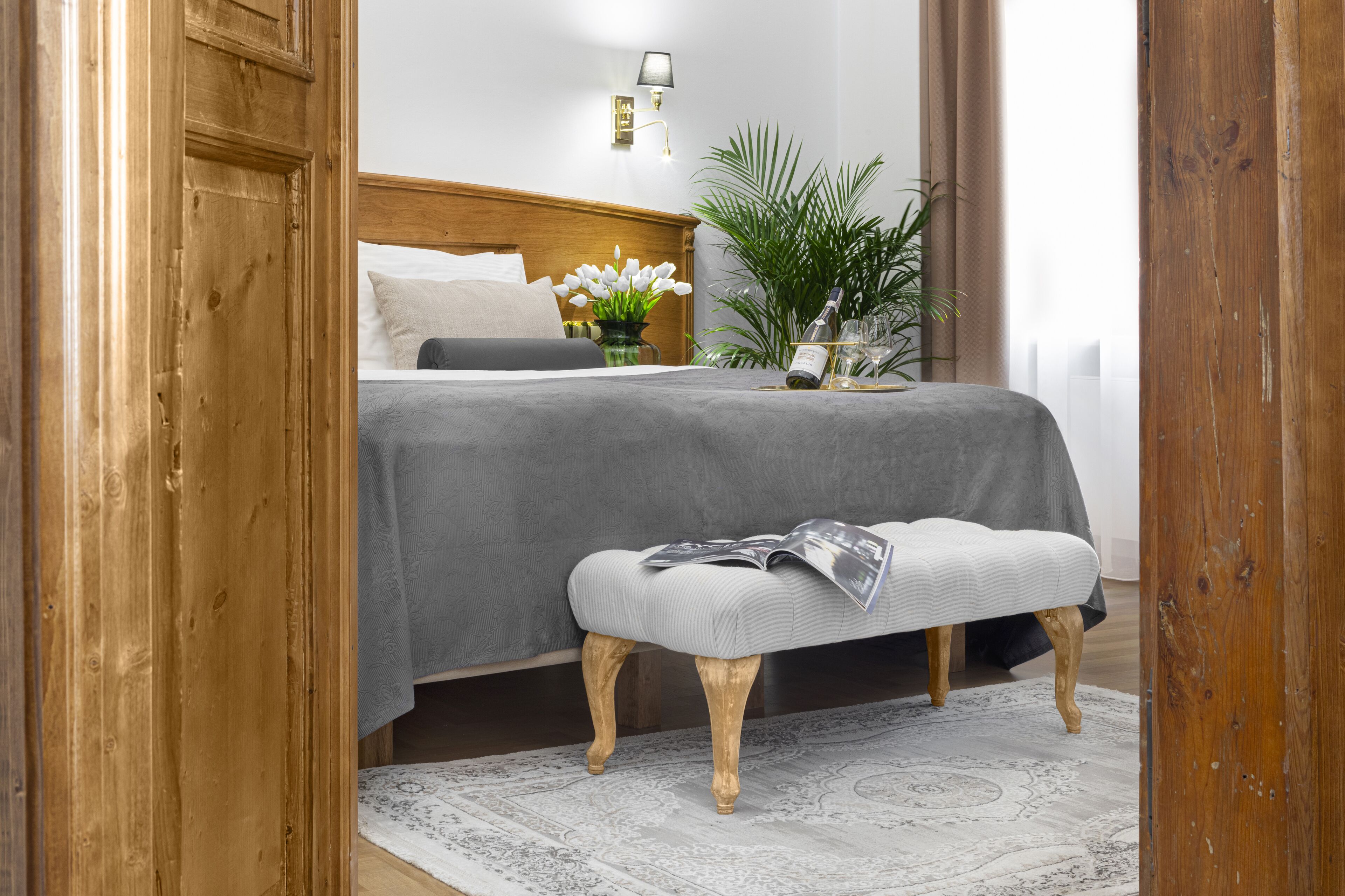 A bedroom with a wooden headboard, a grey bedspread, a bedside table with a vase of white flowers, and a bench with a magazine.