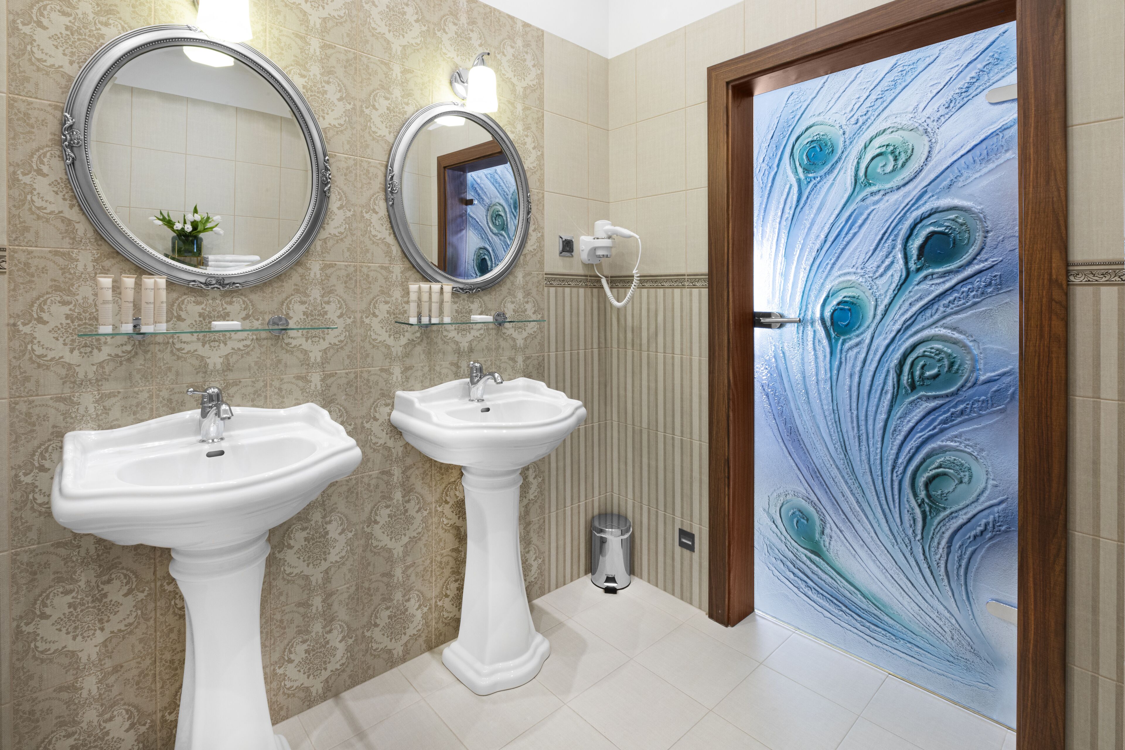 A bathroom with two white pedestal sinks, a round mirror, and a decorative shower door with a peacock feather design.