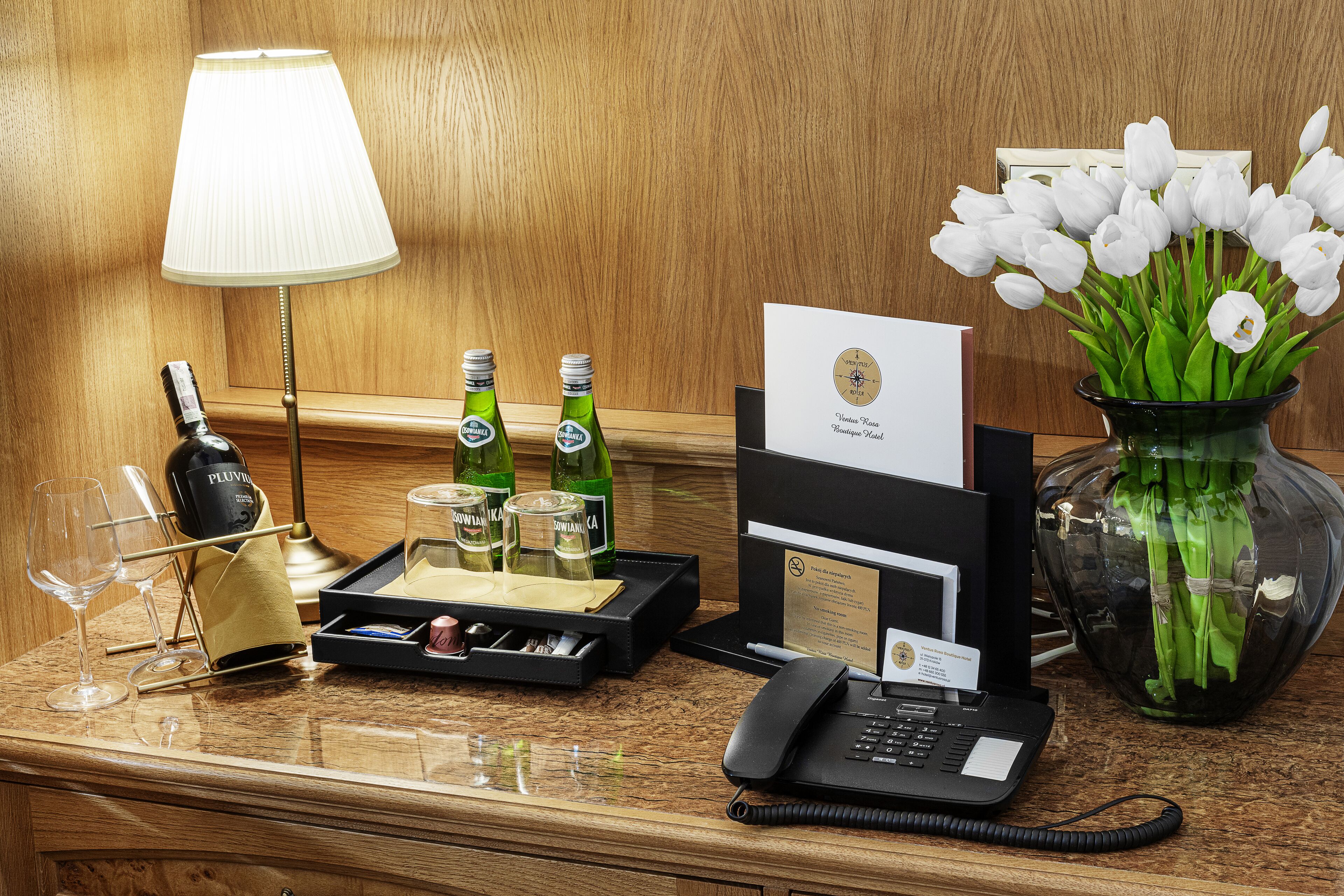 Ein Hotelrezeptionstresen mit einer Lampe, Weinflaschen, Gläsern, einer Blumenvase und einem Telefontablett.