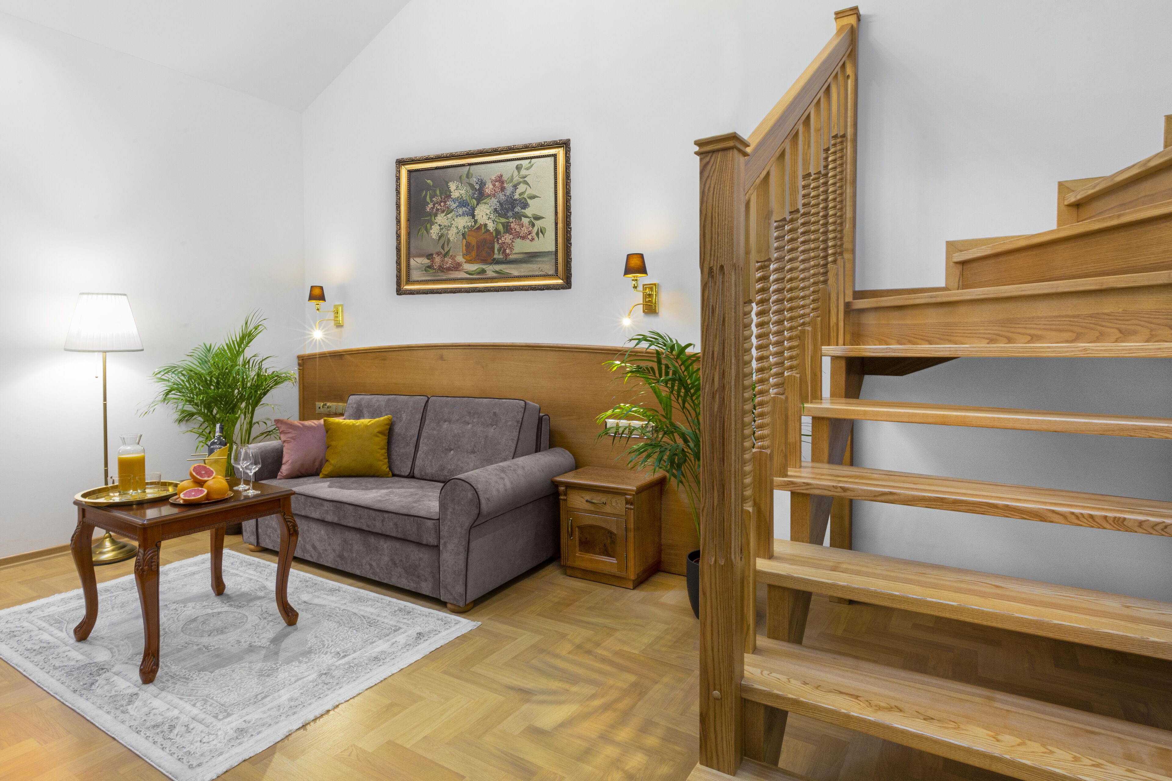 A living room with a grey sofa, a wooden staircase, a painting on the wall, and a small table with a lamp and plants.