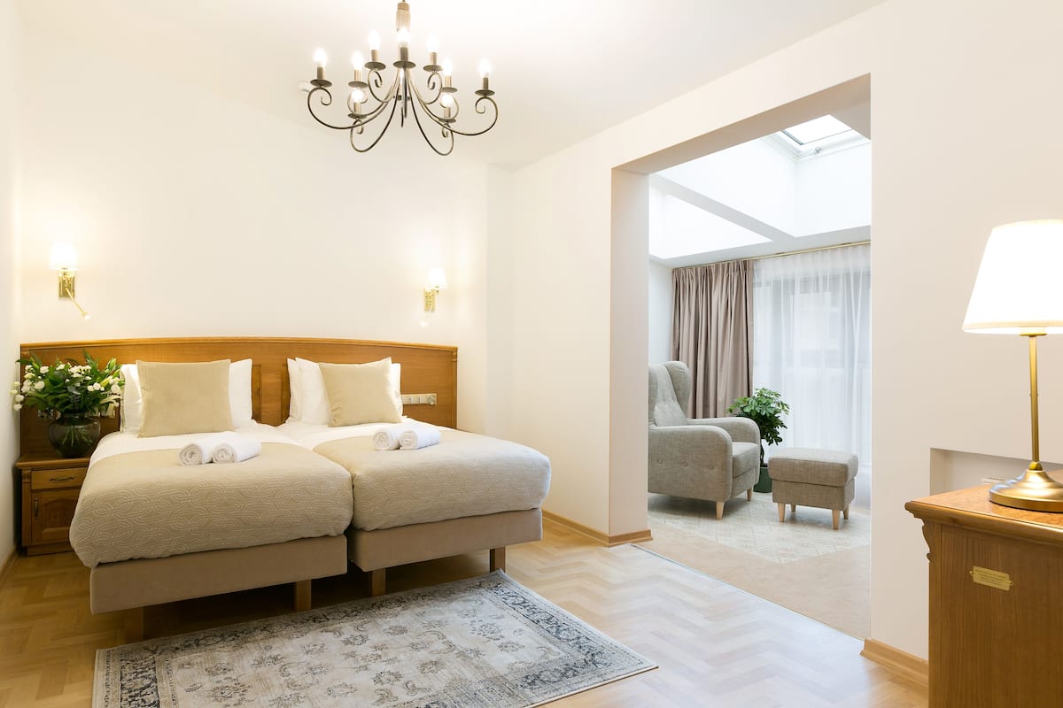 Une chambre d’hôtel avec un lit double, un lustre, un bureau avec une lampe, une chaise et une vue sur le ciel grâce à une lucarne.