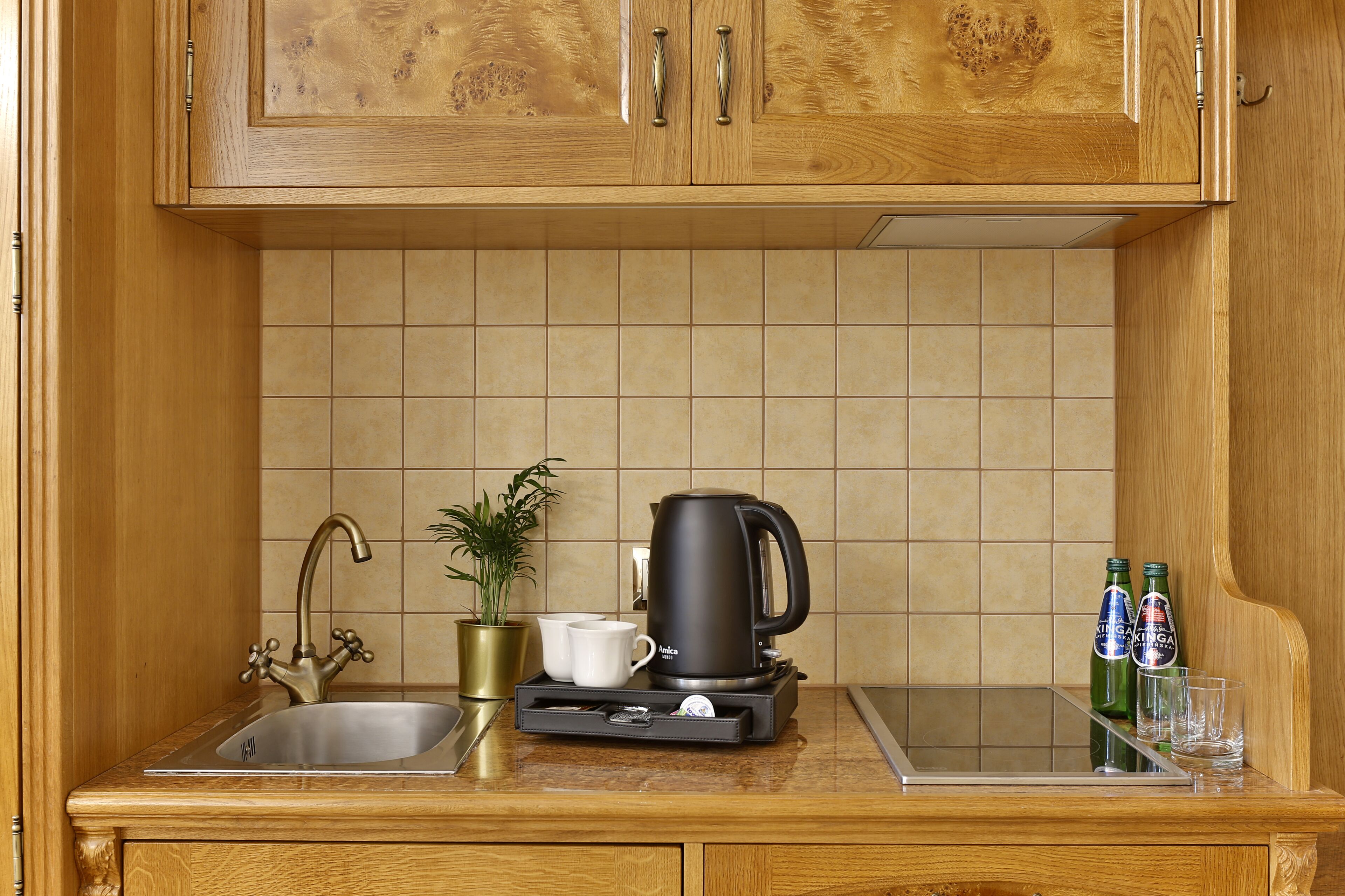 Une cuisine avec des meubles de rangement en bois, un évier en acier inoxydable, une machine à café noire, deux tasses blanches, une plante en pot et deux bouteilles de bière.