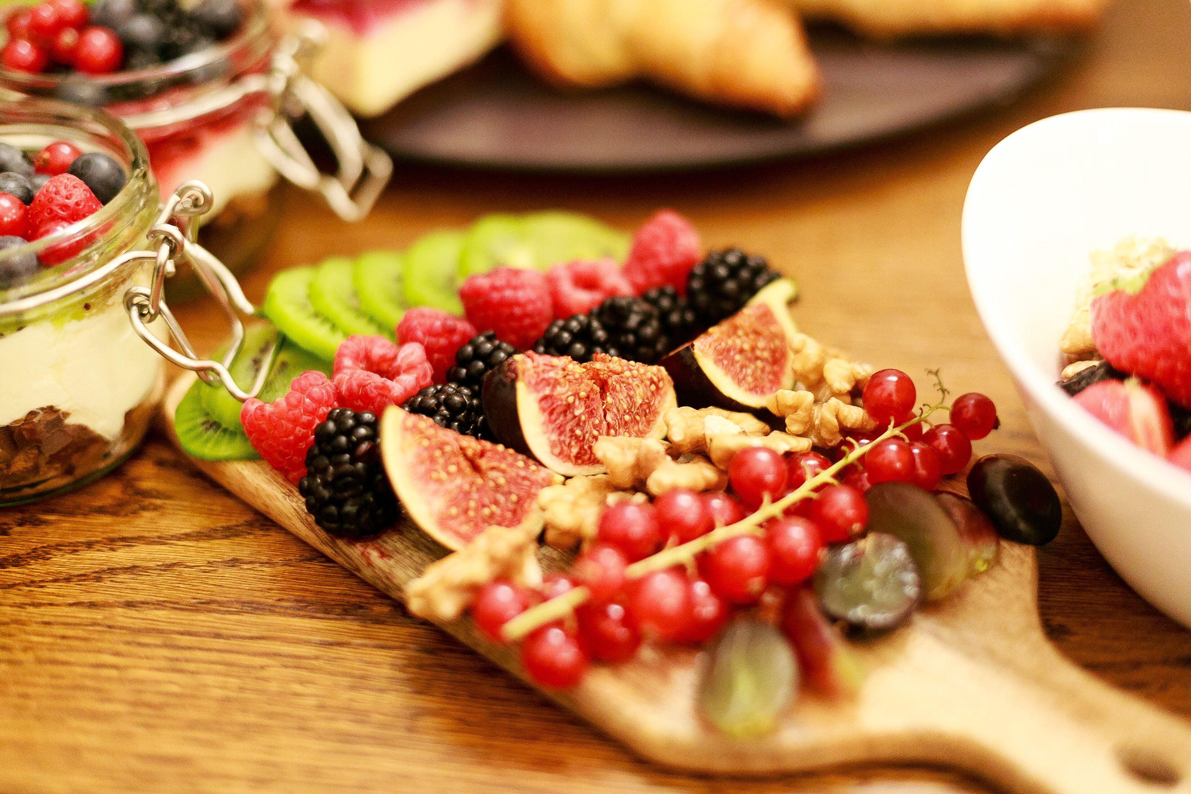 A wooden board with various fruits and nuts, a bowl of yogurt, and a jar of berries.