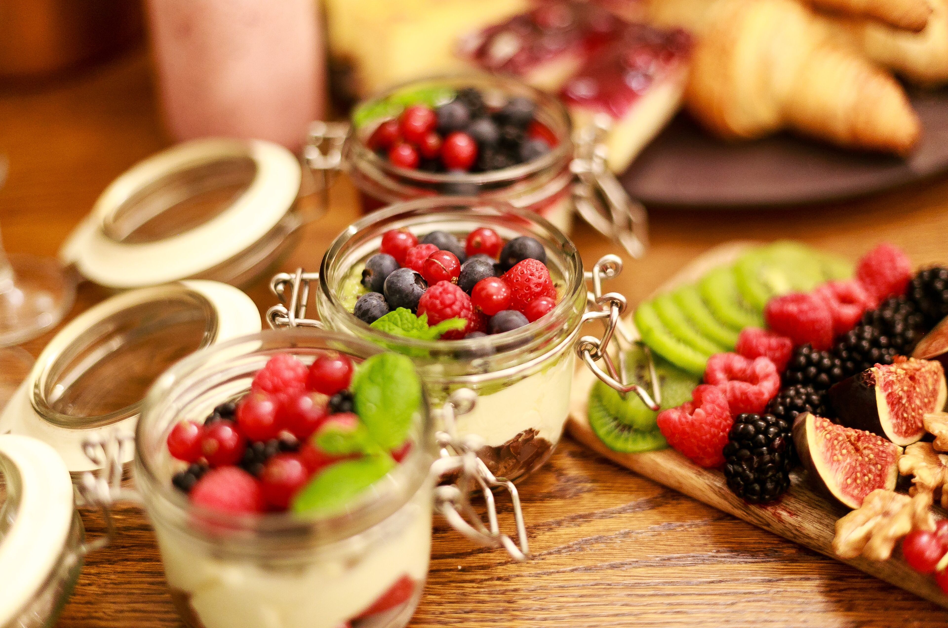 Drie glazen potten met metalen sluitingen gevuld met verschillende bessen en fruit, geplaatst op een houten oppervlak, met een gebakje en ander fruit op de achtergrond.