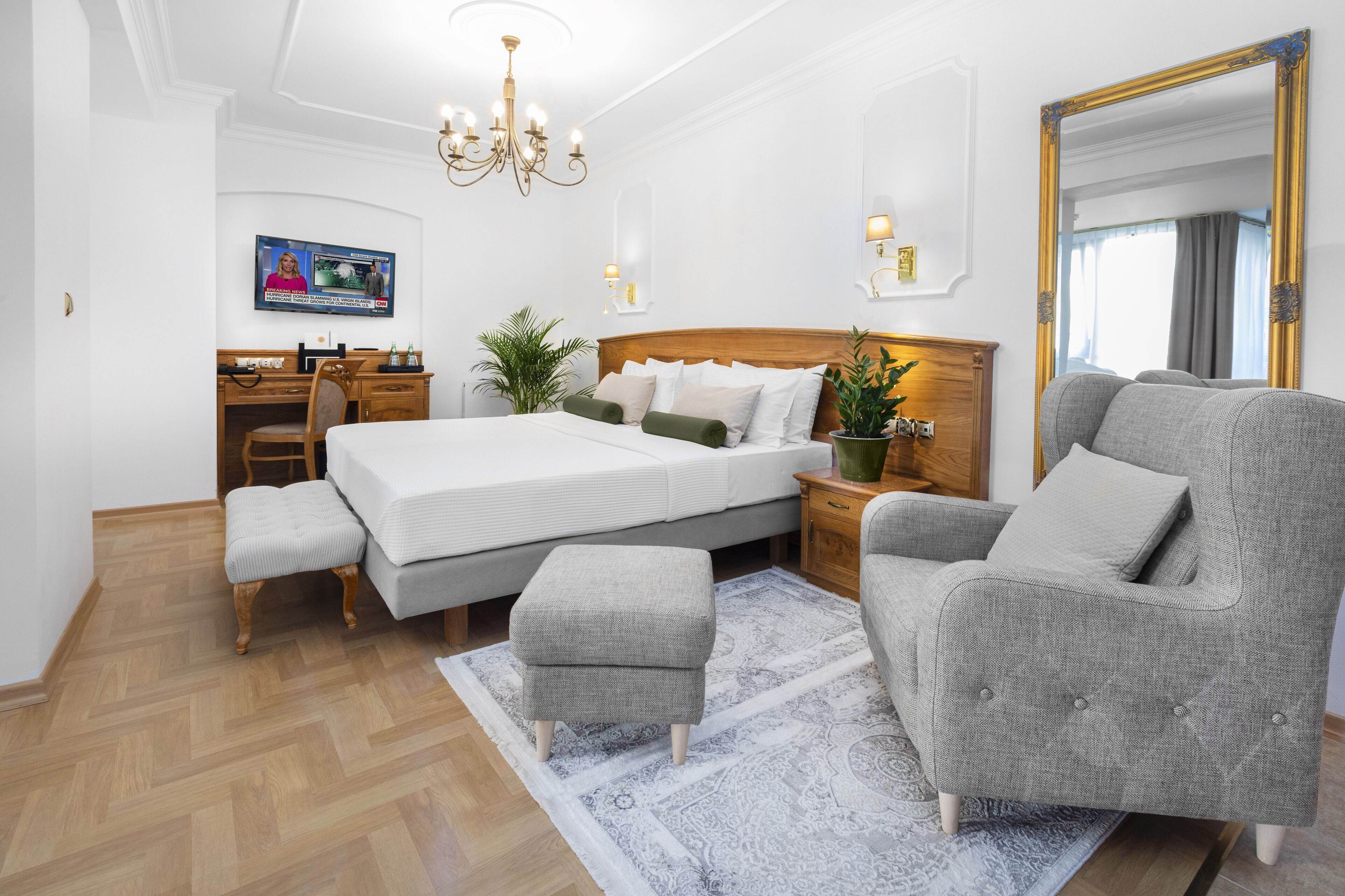Een slaapkamer met een groot bed, een grijze fauteuil, een houten bureau en een televisie aan de muur.