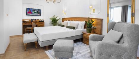 Un dormitorio con una cama grande, un sillón gris, un escritorio de madera y un televisor en la pared.