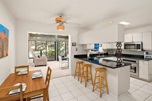 Une cuisine moderne avec des meubles blancs, un îlot central doté de tabourets, un coin repas avec une table et des chaises en bois, et un ventilateur de plafond.