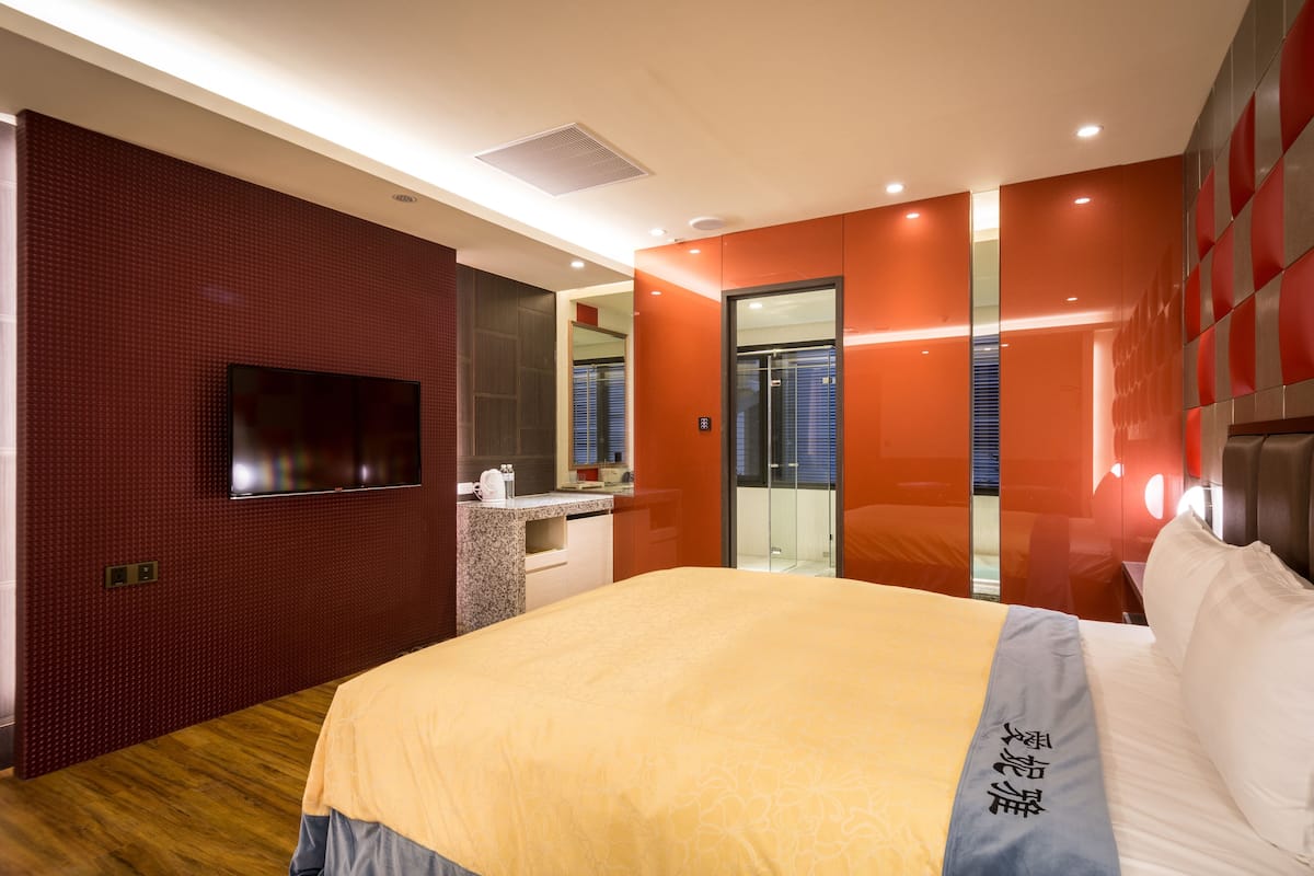 Une chambre d’hôtel moderne dotée d’un grand lit, d’un téléviseur à écran plat fixé au mur, d’une salle de bain avec une porte en verre et d’une décoration aux couleurs rouge et orange.