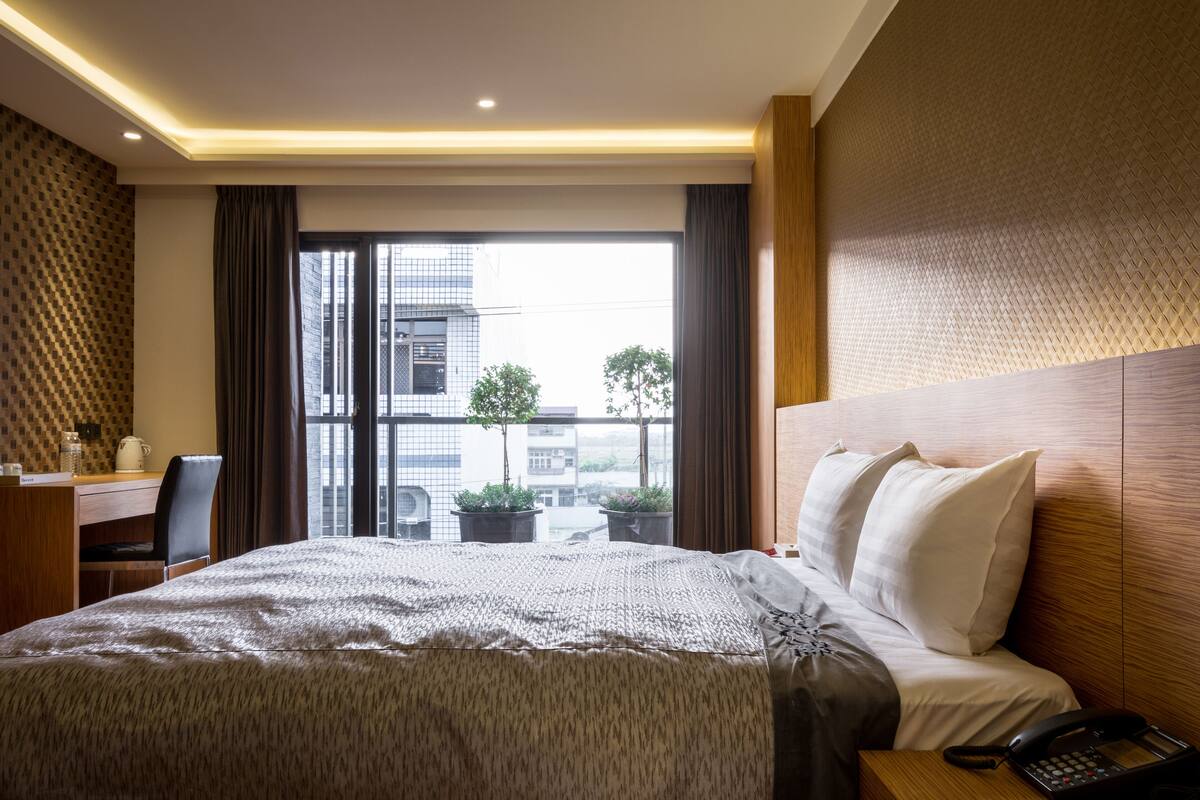 Une chambre d’hôtel avec un grand lit, un bureau avec une chaise, un téléphone et une vue sur les bâtiments par la fenêtre.