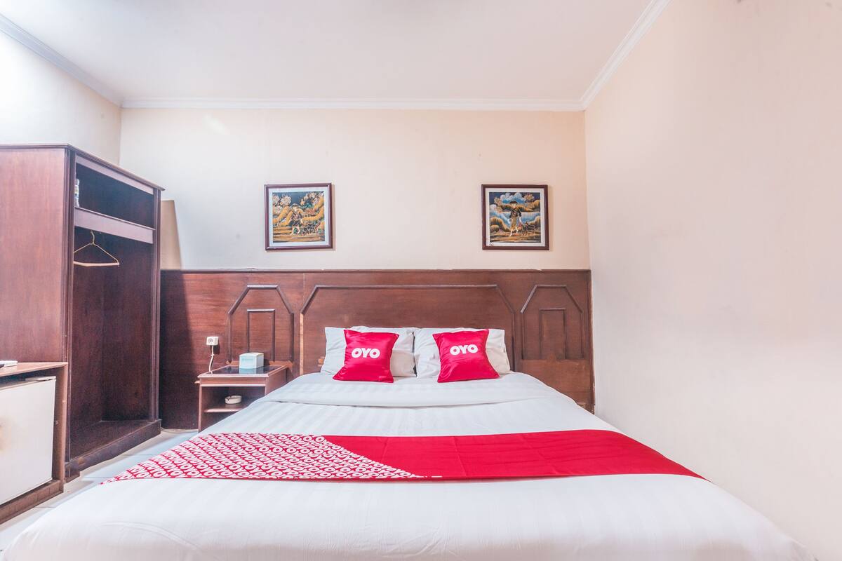 Una cama bien hecha con ropa de cama blanca y roja, dos cuadros en la pared y un armario de madera.