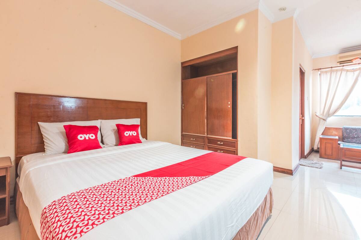 Habitación de hotel con una cama grande, dos almohadas rojas, un armario de madera y vistas al exterior a través de una ventana.