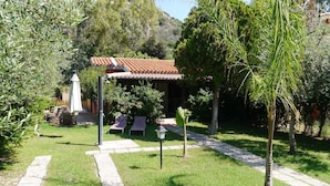 Ein Garten mit einer Pergola, zwei Liegestühlen und einem Weg, der zu einem Gebäude mit einem Ziegeldach führt.