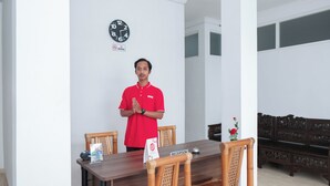 A man in a red shirt standing in a dining area with a wooden table, chairs, and a clock on the wall. - Oyo 1454 111 Residence (Batu)