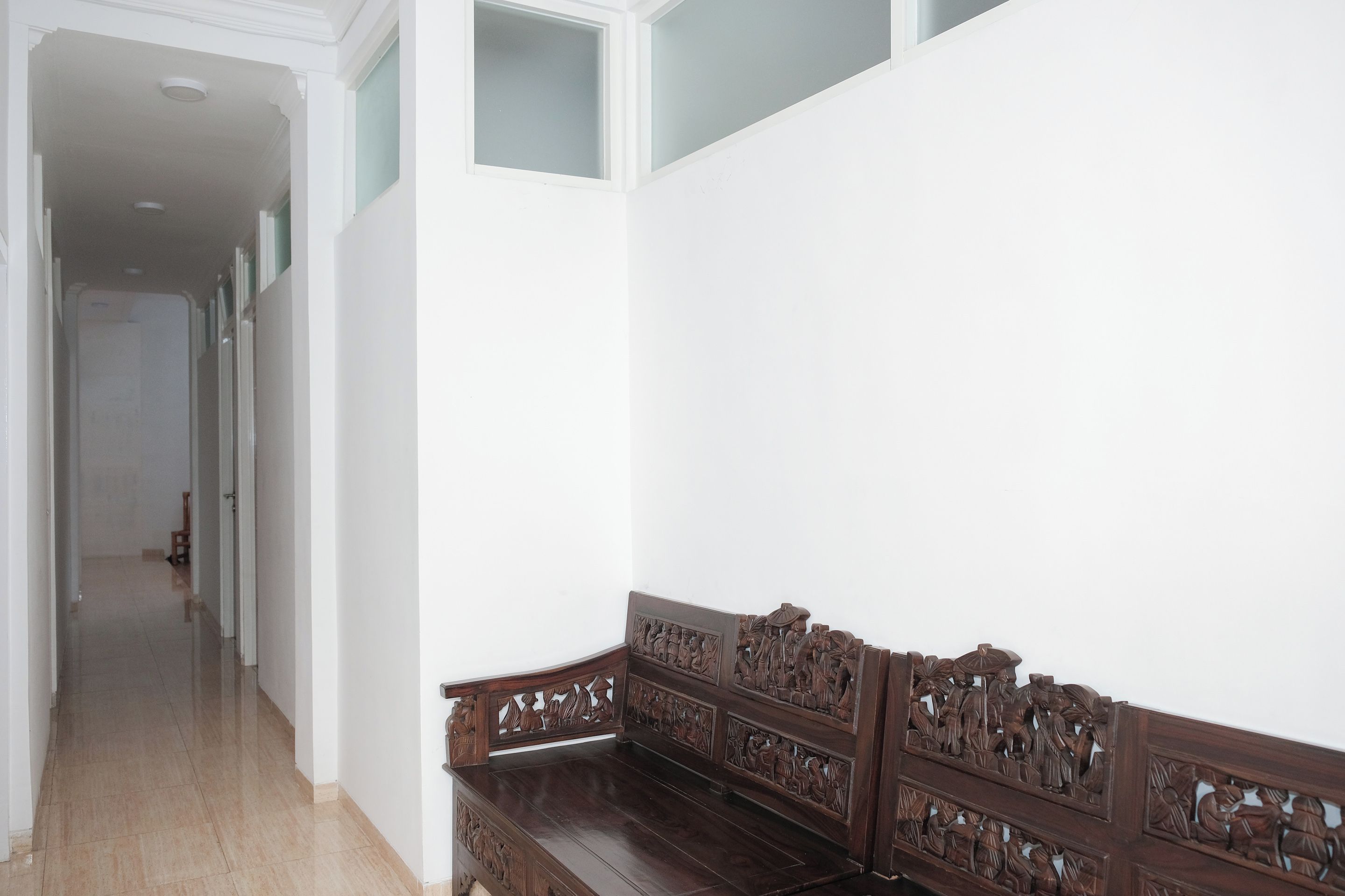 A hallway with a wooden bench featuring intricate carvings, a tiled floor, and a white wall with a window.