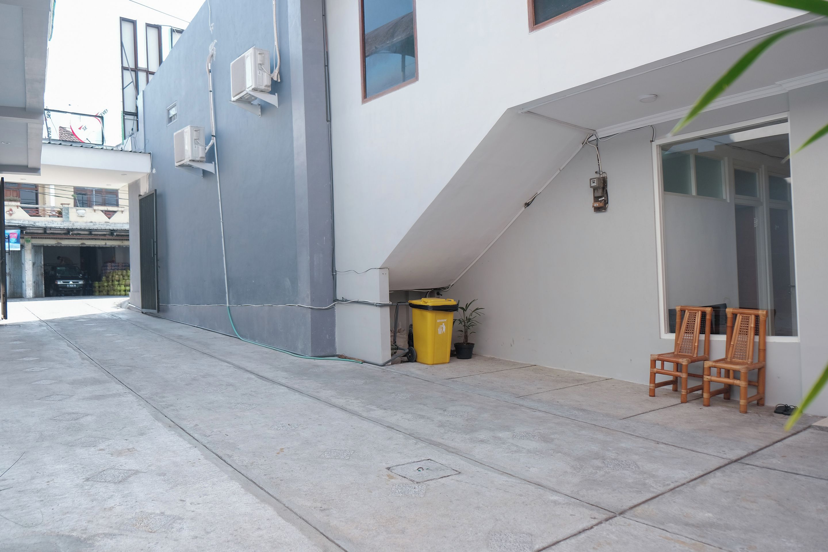 A building with a grey exterior, a visible window, and a door. There is a yellow trash bin and a potted plant near the entrance.