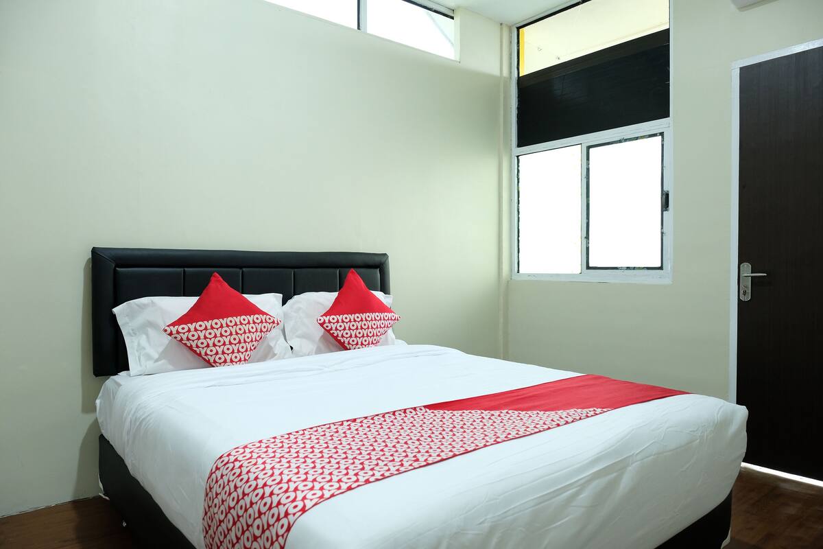 Una cama bien hecha con ropa de cama blanca y roja, un cabecero negro y una ventana con persianas.