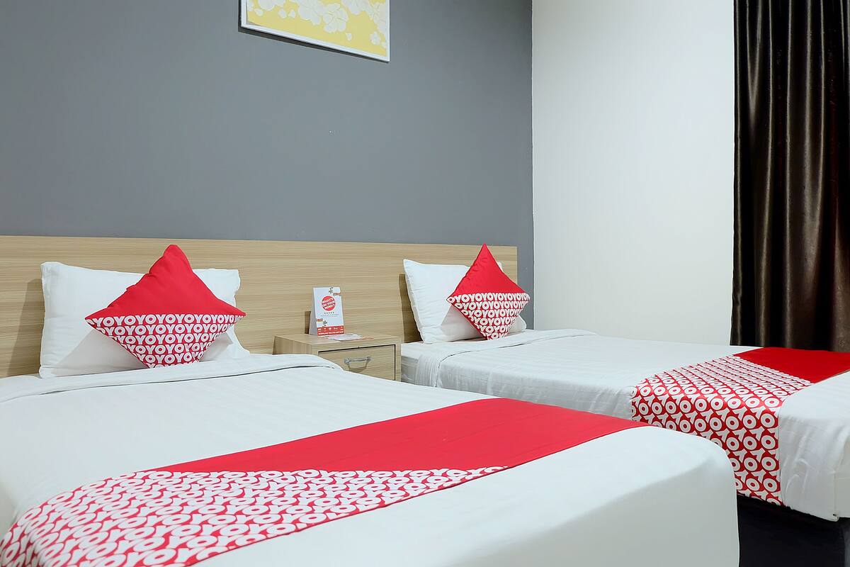 Habitación de hotel con dos camas, cada una con ropa de cama blanca y detalles rojos, un cabecero de madera y una mesita de noche con lámpara.