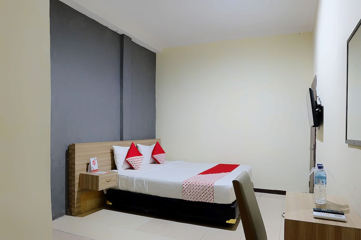 Una cama bien hecha con una colcha blanca y roja, una mesita de noche de madera con una lámpara, y una botella de agua sobre una pequeña mesa junto a la cama.
