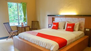 A hotel room with a large bed, two pillows with the brand name "OYO," a wooden headboard, a small table with a chair, and a window with a view of greenery. - Hotel O Jatimas (Merawang)