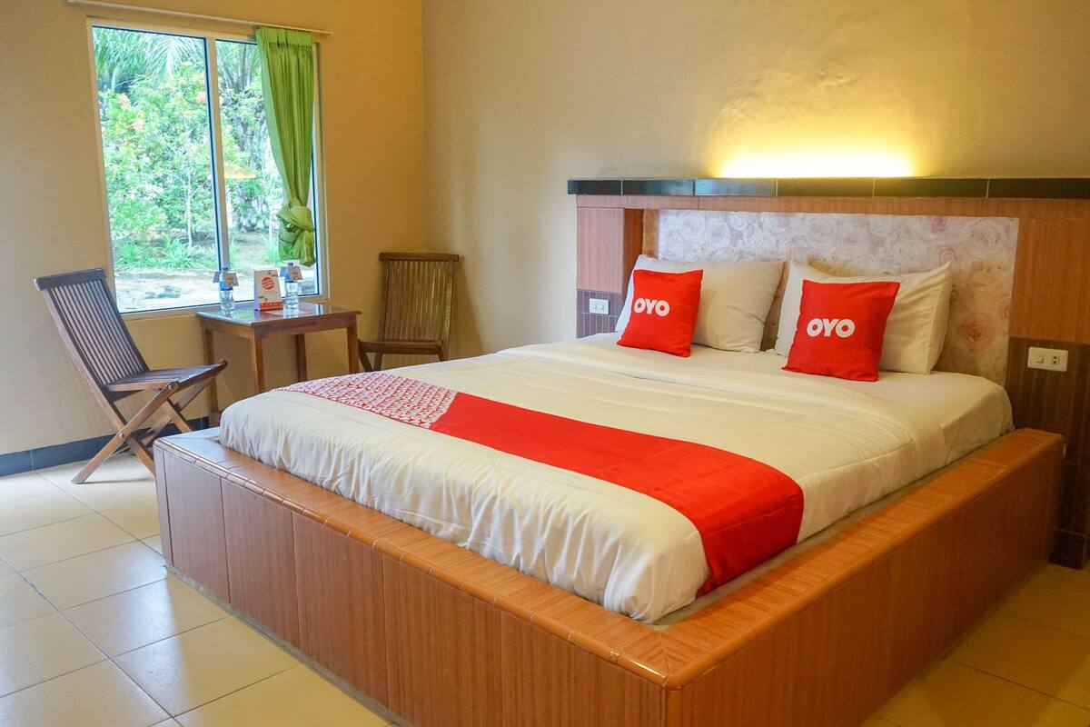 Ein Hotelzimmer mit einem großen Bett, zwei Kissen mit dem Schriftzug „OYO“, einem hölzernen Kopfteil, einem kleinen Tisch mit Stuhl und einem Fenster mit Blick auf Grünflächen.