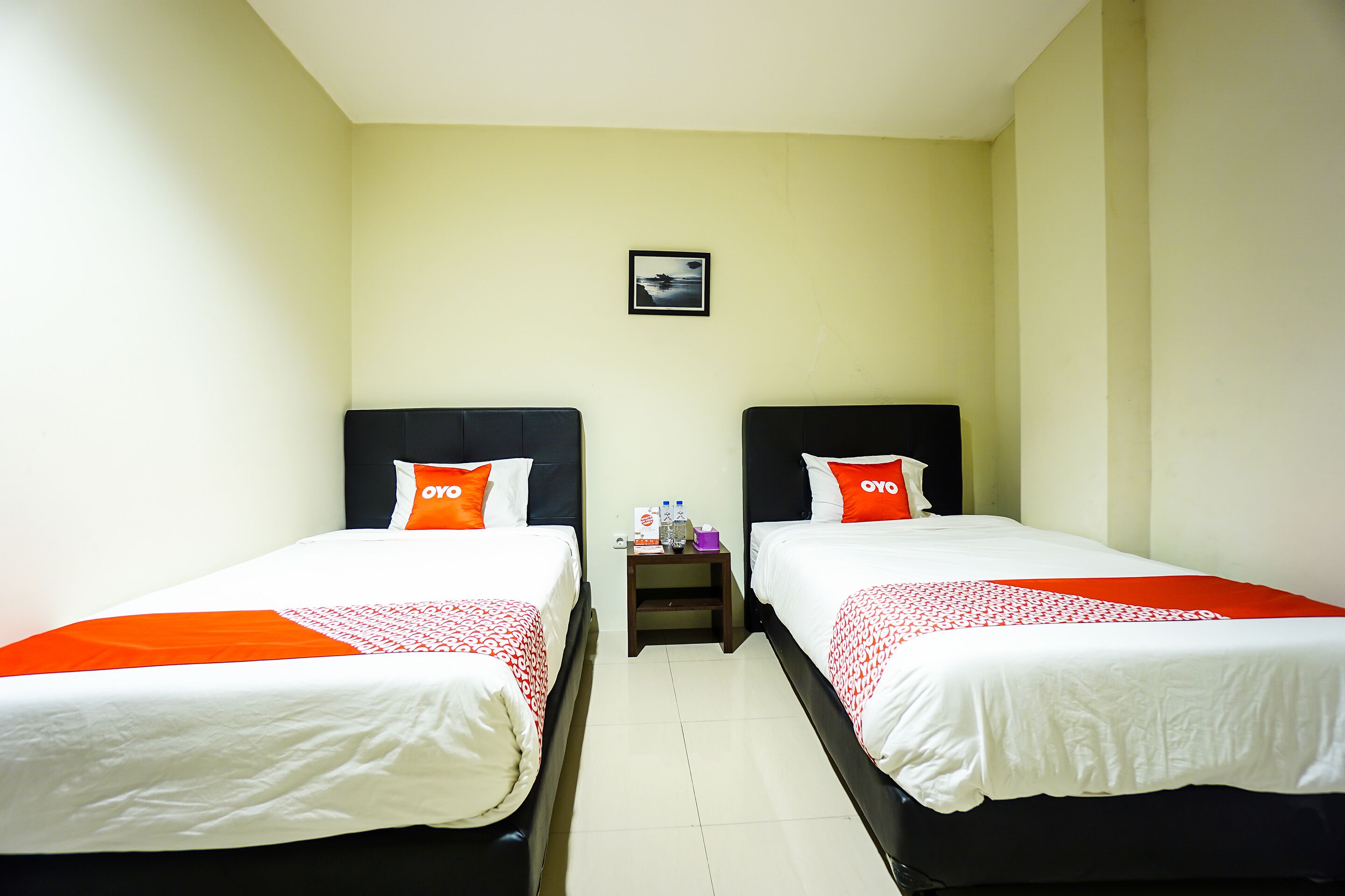 A hotel room with two beds, each with white bedding and orange pillows, a small bedside table with a water bottle and a cup, and a framed picture on the wall.
