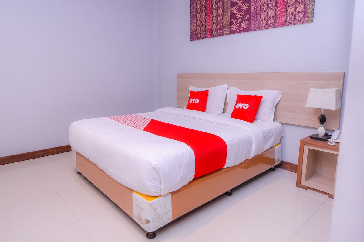 Een netjes opgemaakt bed met een wit-rode sprei, twee rode kussens met 'OYO'-branding, een nachtkastje met een lamp, en een wand met een patroonwandkleed.