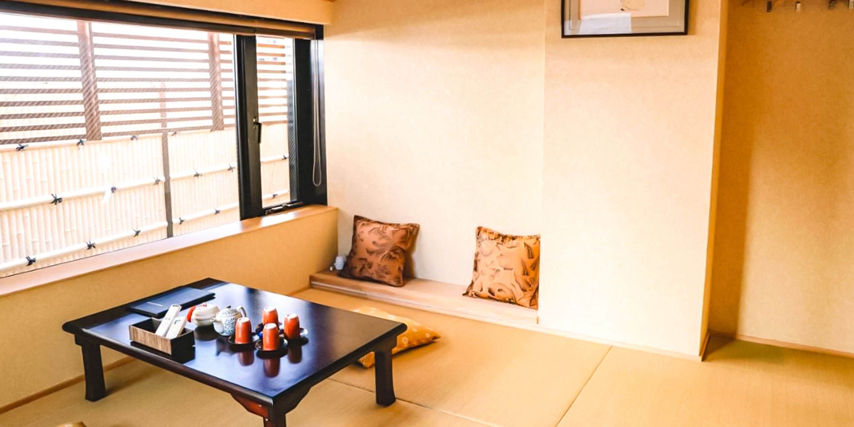 Une pièce avec un sol recouvert de tatamis, une petite table sur laquelle sont posés divers objets, une fenêtre avec des stores et un coussin sur un banc bas.