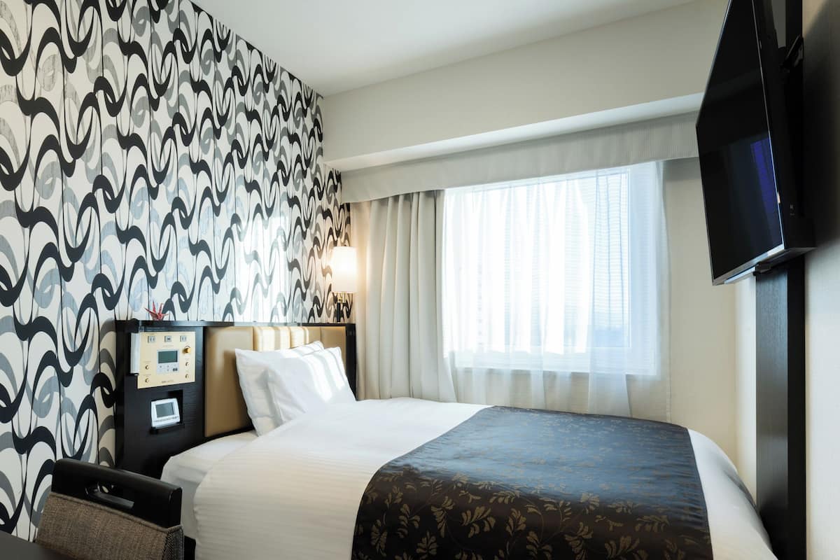 A hotel room with a large bed, a bedside table, a TV mounted on the wall, and a window with curtains.