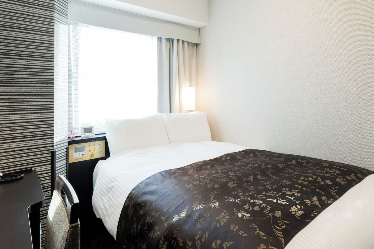A hotel room with a bed, a desk, a chair, a lamp, and a window with curtains.