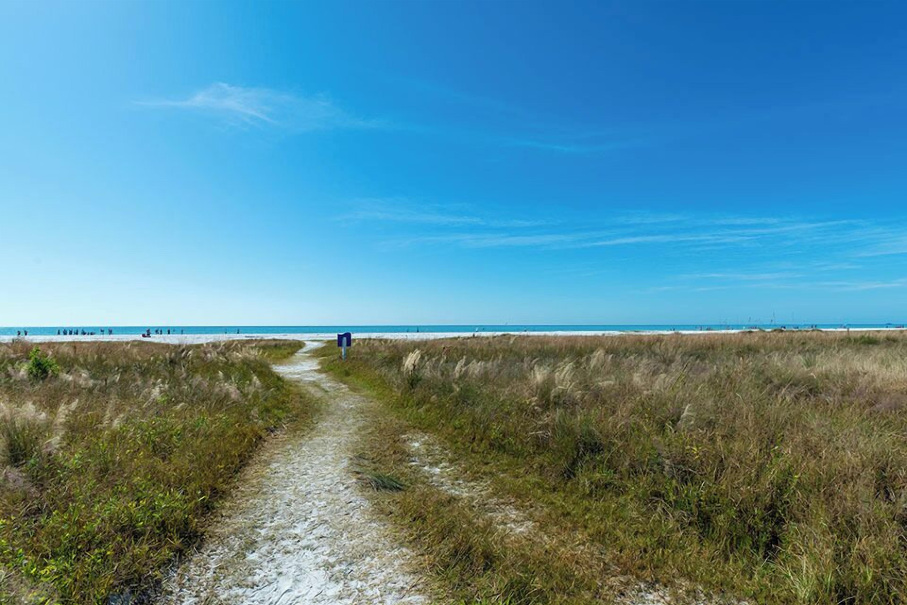 Ein sandiger Pfad führt zu einem Strand, der von blauem Himmel überspanned wird.