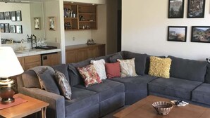 A living room with a sofa, coffee table, and a lamp. There is a kitchen area with cabinets and a mirror.