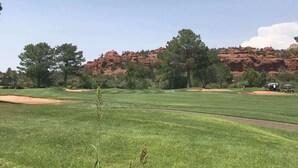 A golf course with a clear sky, green grass, trees, and red rock formations in the background.