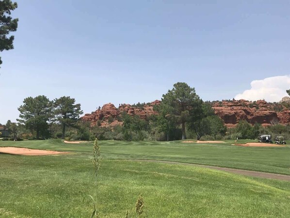A golf course with a clear sky, green grass, trees, and red rock formations in the background.