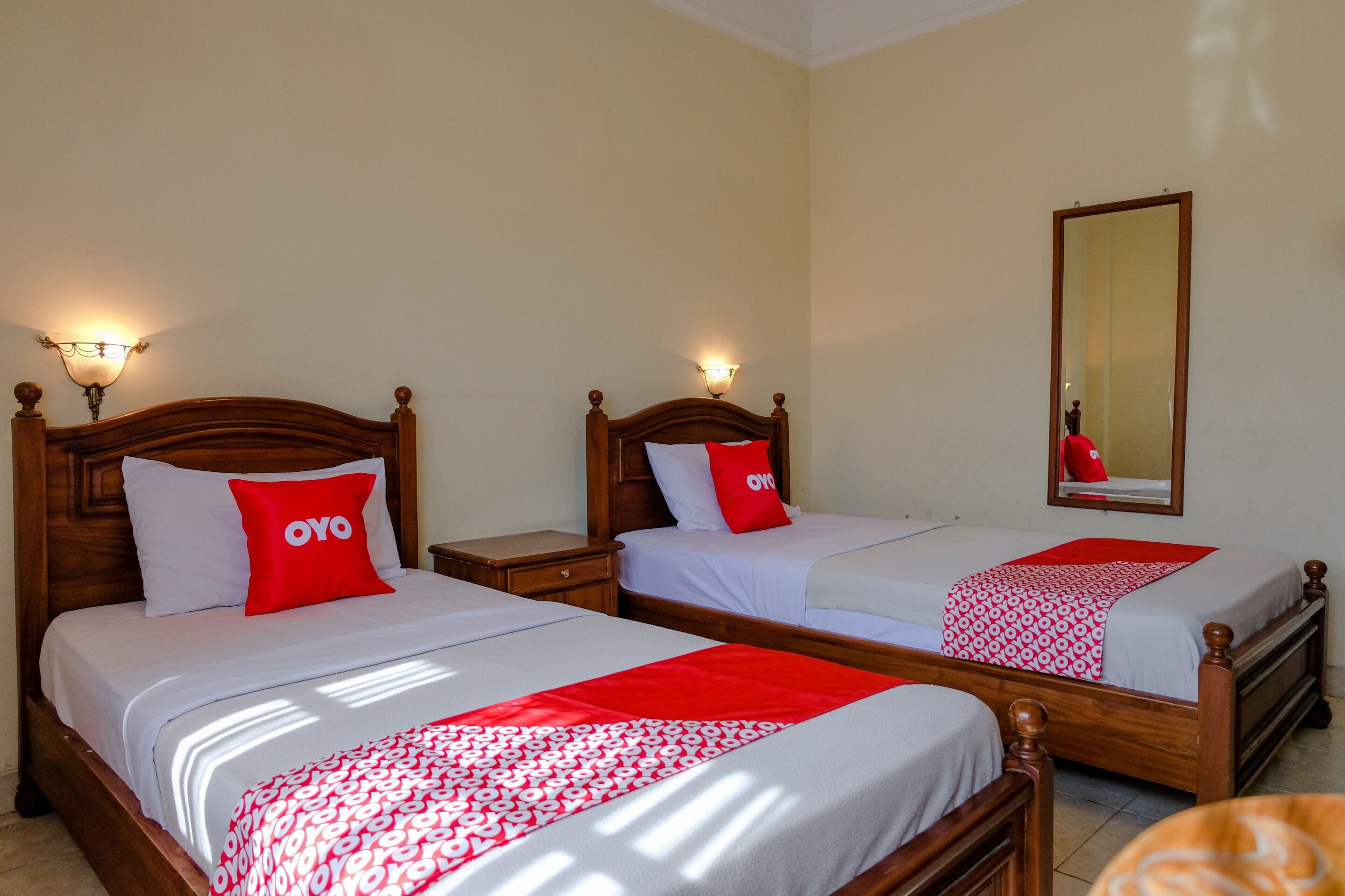 Habitación de hotel con dos camas, cada una con cabecero de madera, ropa de cama blanca y almohadas rojas. Incluye lámparas de noche, un espejo y una manta estampada en rojo y blanco.