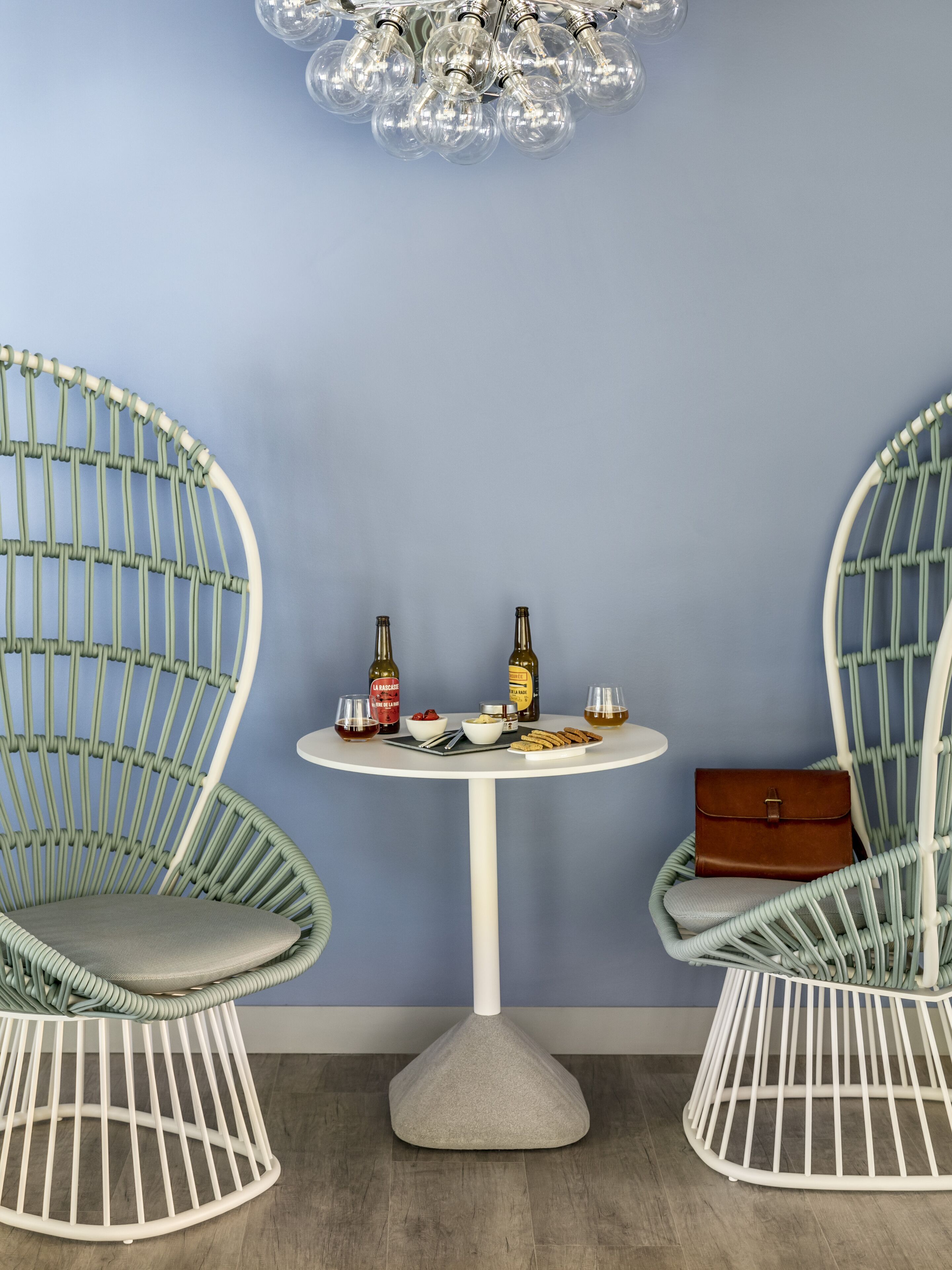 A room with two wicker chairs, a small round table with drinks and snacks, and a brown leather bag on the table.