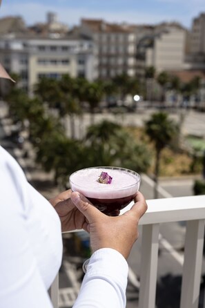 A person holding a drink with a purple hue, possibly a cocktail, on a balcony with a cityscape in the background.