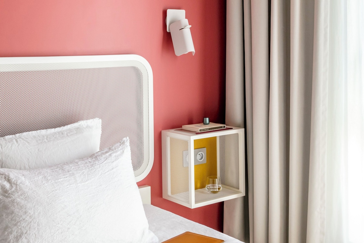 A neatly made bed with white linens, a white headboard, and a bedside table with a lamp and a small shelf.
