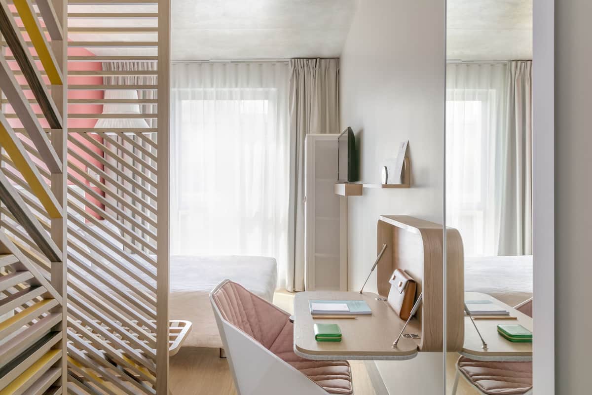 Une chambre d’hôtel moderne équipée d’un lit, d’un bureau avec une chaise, d’un miroir et d’une fenêtre avec des rideaux.