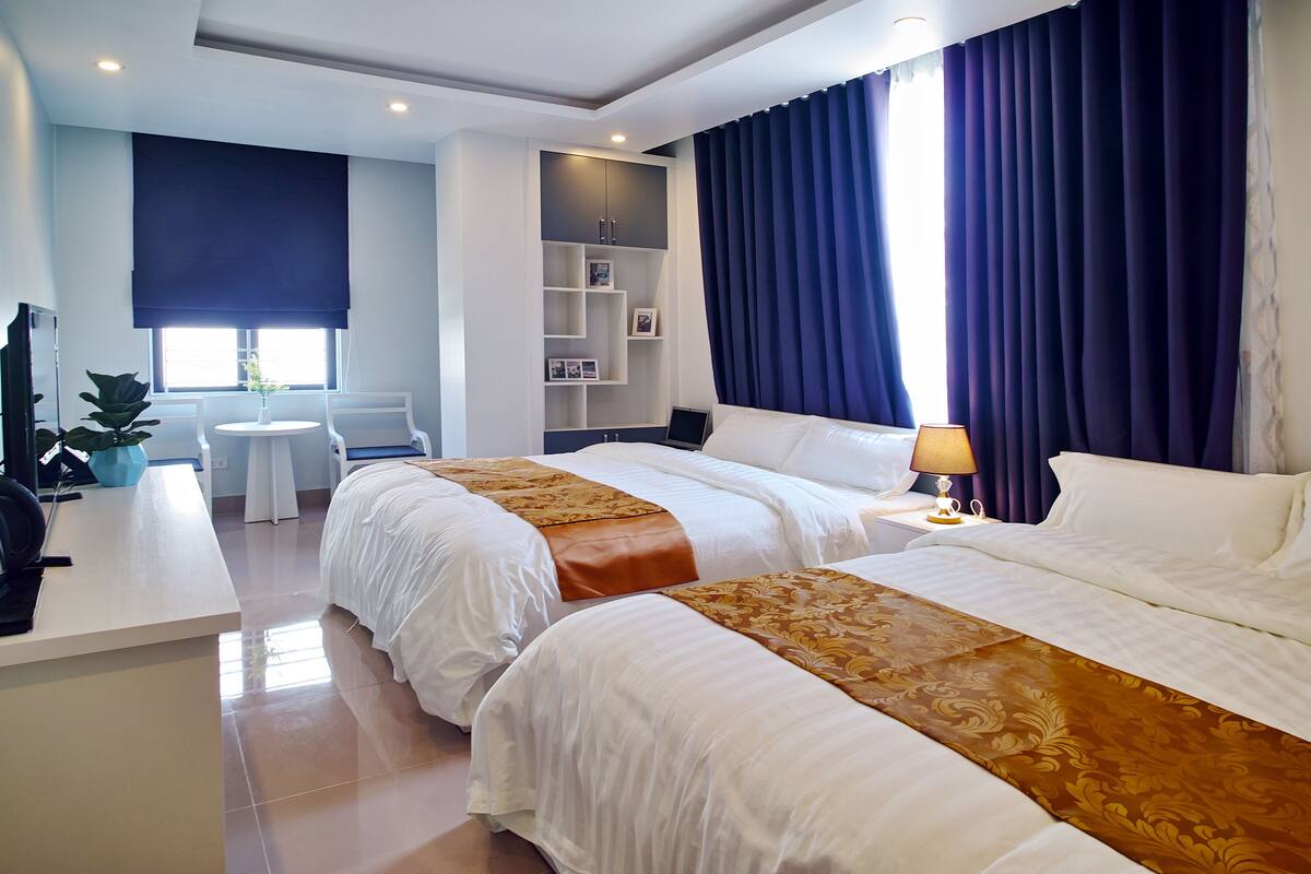 A hotel room with two beds, a desk, a chair, a small table, and a TV.