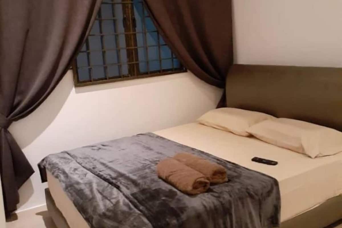 Una cama individual con una manta gris y dos toallas marrones, una ventana con barrotes y una unidad de aire acondicionado sobre la ventana.