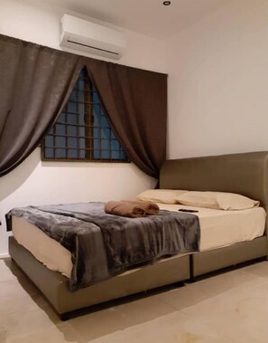 A single bed with a grey headboard and beige bedding, a brown towel on the bed, and an air conditioning unit above a window with brown curtains.