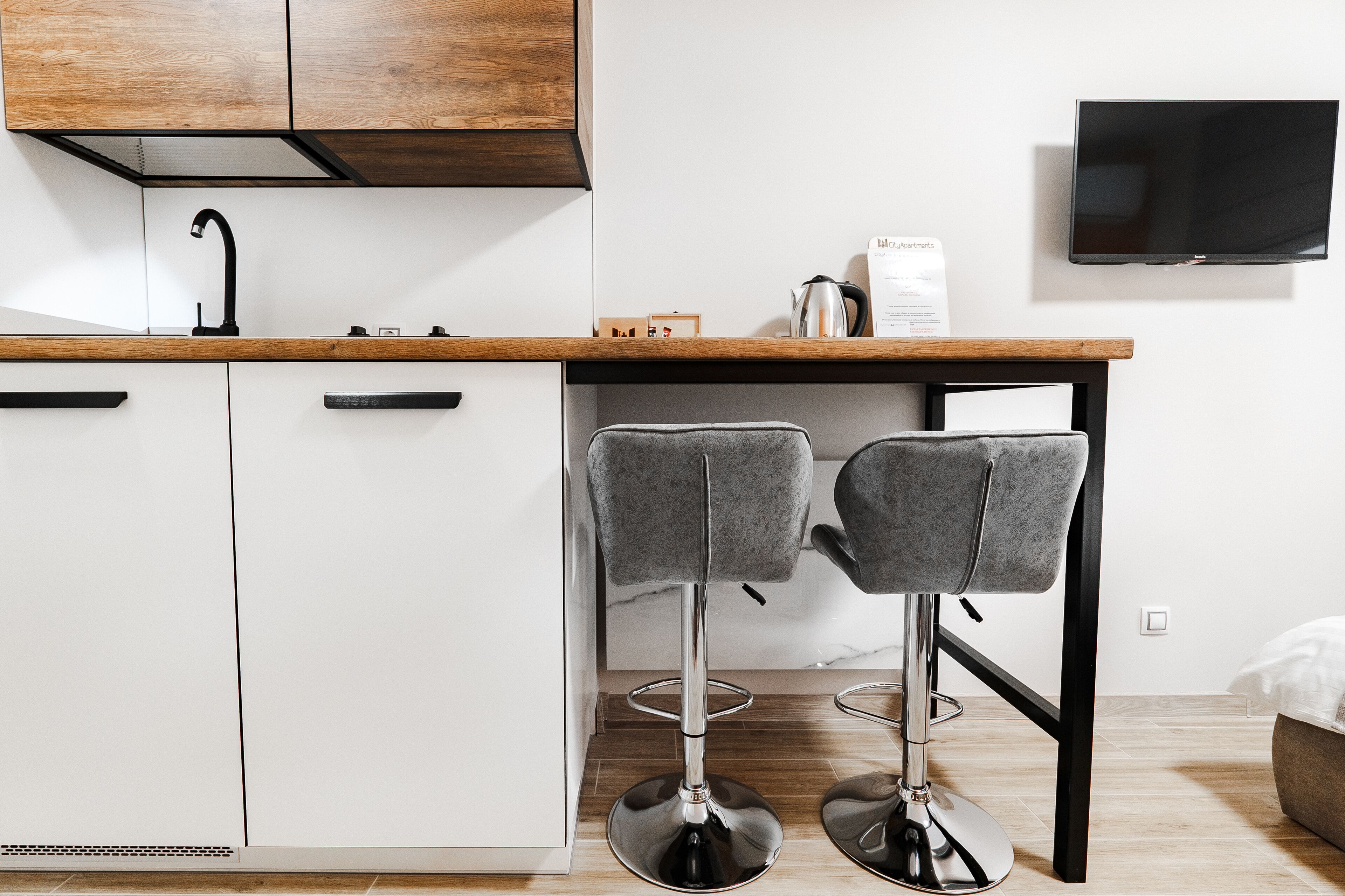 A modern kitchen with white cabinets, a wooden countertop, a black faucet, and two grey bar stools. A flat-screen TV is mounted on the wall above the counter.