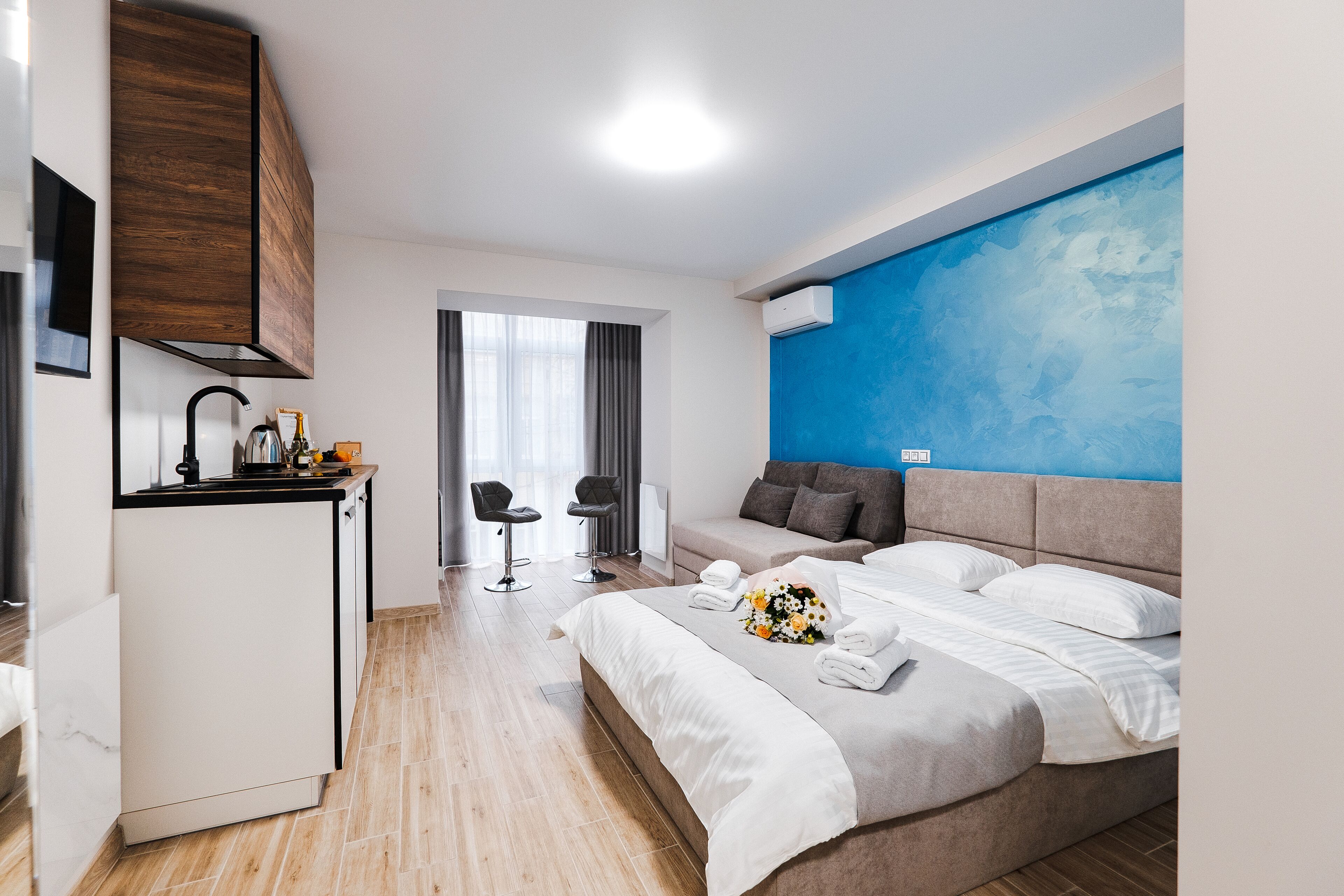 Ein modernes Hotelzimmer mit einem Bett, einer kleinen Küchenzeile, einer Sitzecke mit zwei Stühlen und einem großen Fenster.