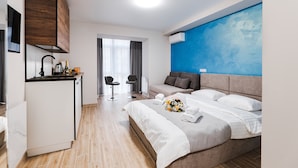 Une chambre d’hôtel moderne comprenant un lit, une petite kitchenette, un coin salon avec deux fauteuils et une grande fenêtre.