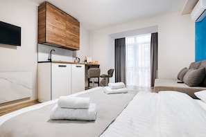 Moderni hotellihuone, jossa on sänky, sohva, työpöytä tuolilla, pieni keittiönurkkaus ja suuri ikkuna, jossa on verhot.