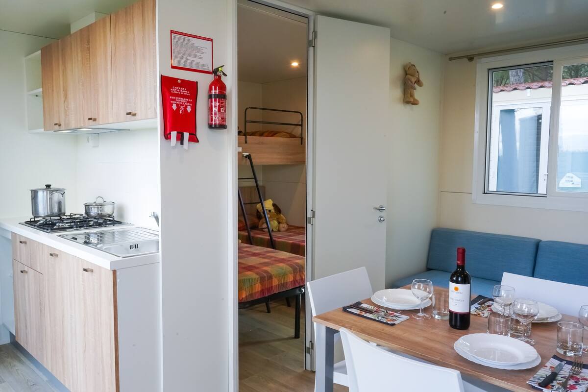 A compact kitchen with a gas stove, sink, and wooden cabinets. A dining area with a table set for two, a sofa, and a window with a view.
