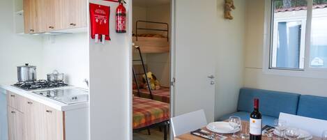 A compact kitchen with a gas stove, sink, and wooden cabinets. A dining area with a table set for two, a sofa, and a window with a view.