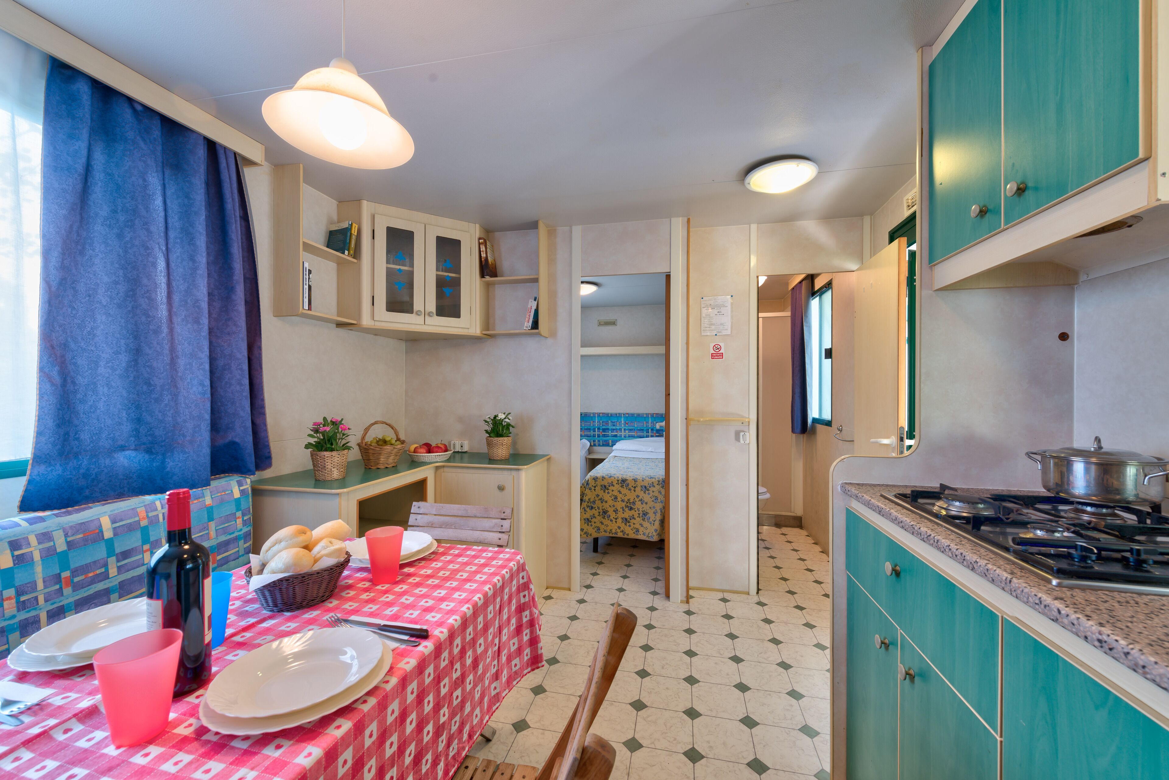 A compact kitchen with a dining area, a bed in the background, and a small window with curtains.