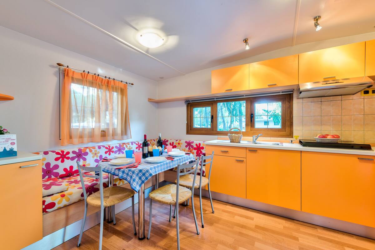 A compact kitchen with a dining area, featuring a table set for four, a window with curtains, and orange cabinetry.