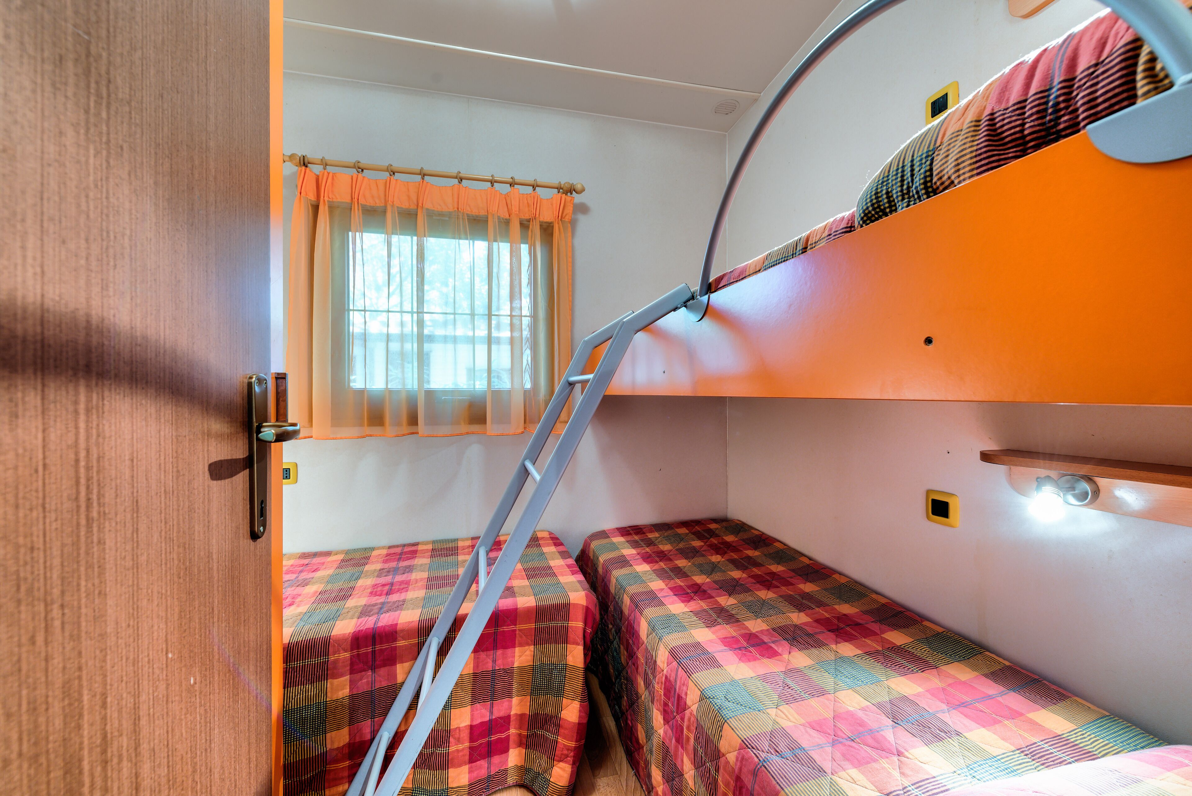 Interior de uma RV compacta com duas camas, uma janela com cortinas e uma escada.